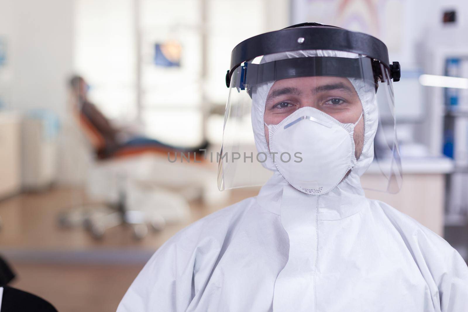 Portrait of tired doctor in dental office looking on camera wearing overall and face shield sitting on chair in waiting room clinic. Concept of new normal dentist visit in coronavirus outbreak.
