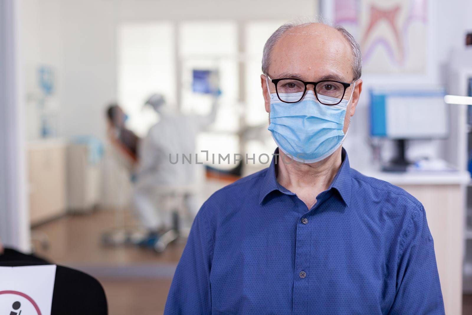 Portrait of elderly man in dental office looking on camera wearing face mask sitting on chair in waiting room stomatological clinic. Concept of new normal dentist visit in coronavirus outbreak.