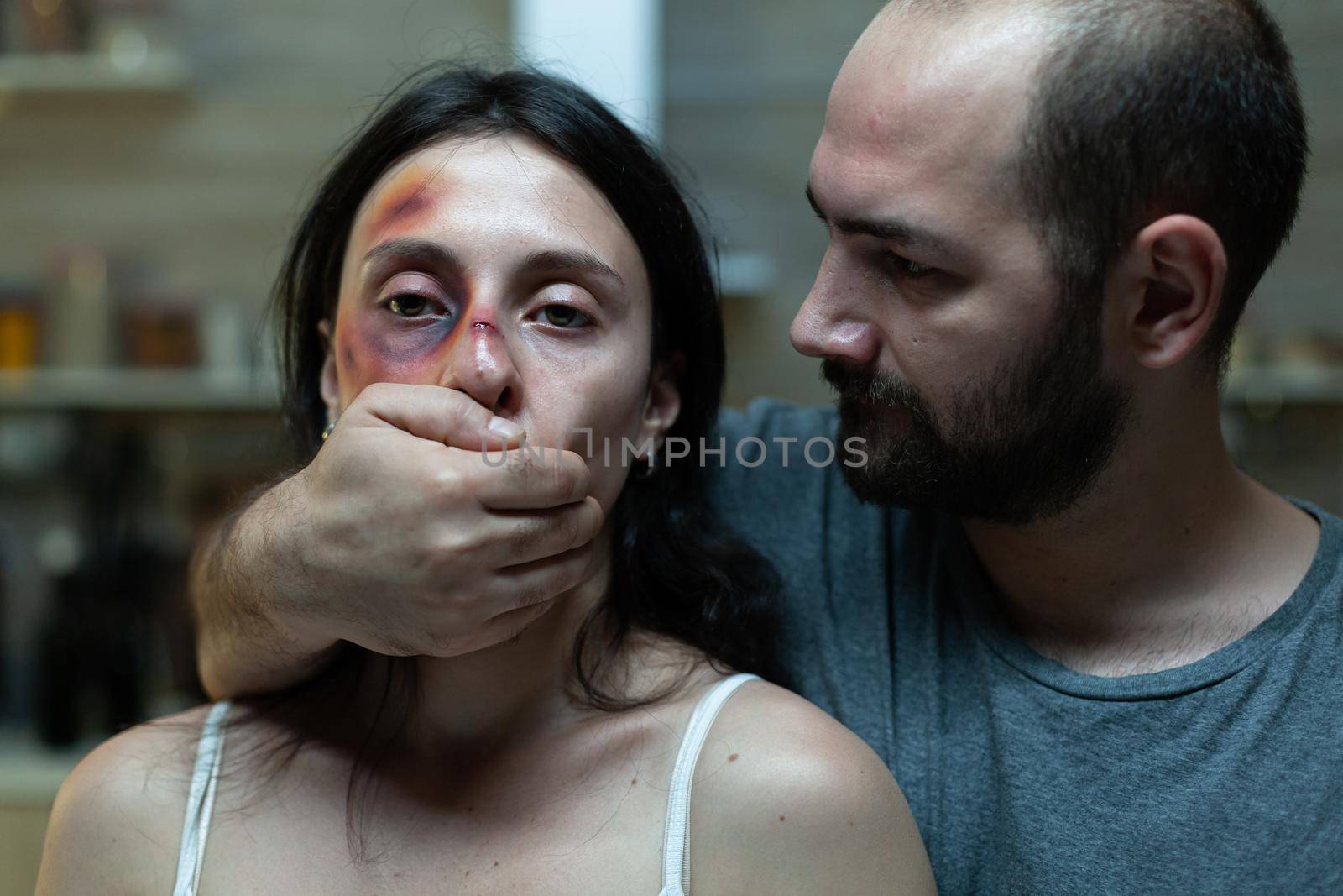 Criminal adult abusing afraid woman with bruises on face while holding hand on mouth for silence. Close up of couple with domestic issues and anger problems. Caucasian people violence