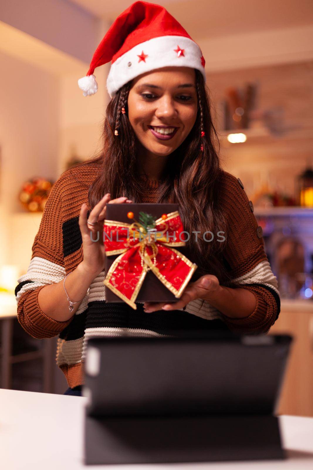 Young woman wearing santa hat using video call technology on tablet at home with ornaments, decorations and tree in kitchen. Caucasian person showing gift with ribbon for christmas party