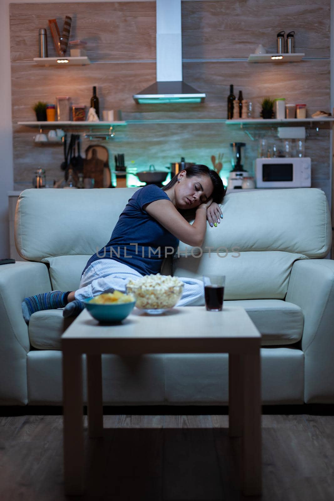 Tired woman closing eyes while watching movie at night. Tired exhausted lonely sleepy housewife in pajamas sleeping in front of television sitting on cozy couch in living room at home.