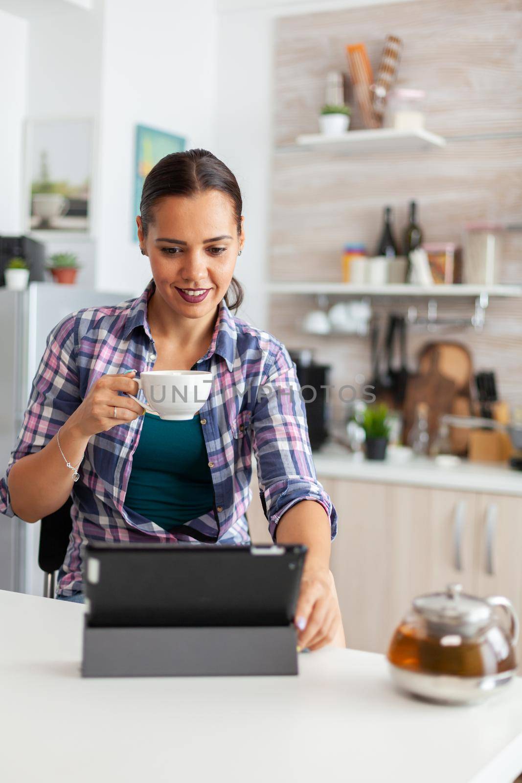 Woman smiling using tablet pc in the morning and enjoying a cup of hot green tea. Working from home using device with internet technology, typing, on gadget during breakfast.