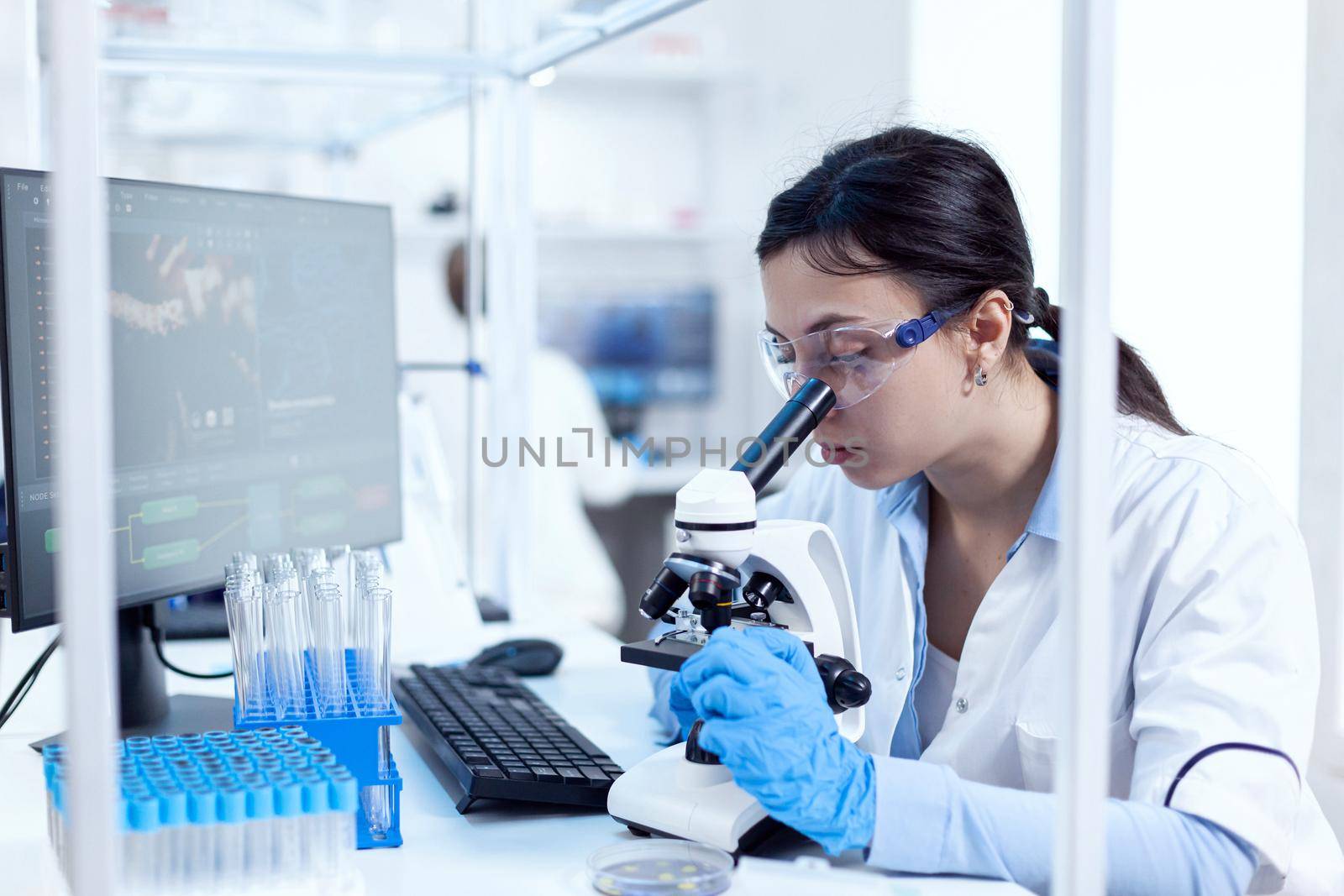 Microbiologist in research laboratory doing scientific experiments looking through microscop. Chemist wearing lab coat using modern technology during scientific experiment in sterile environment.