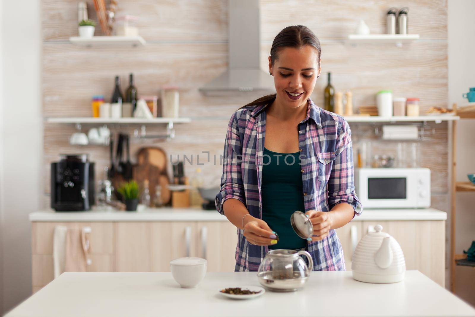 Caucasian woman using aromatic herbs to prepare hot tea in the morning. Preparing tea in a modern kitchen sitting near the table. Putting with hands, healthy herbal in pot.