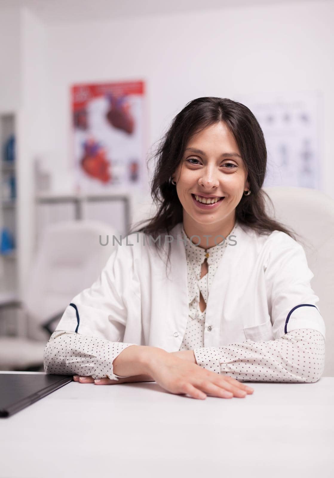 Portrait of smiling woman physician at desk in hospital office wearing white coat looking at the camera.
