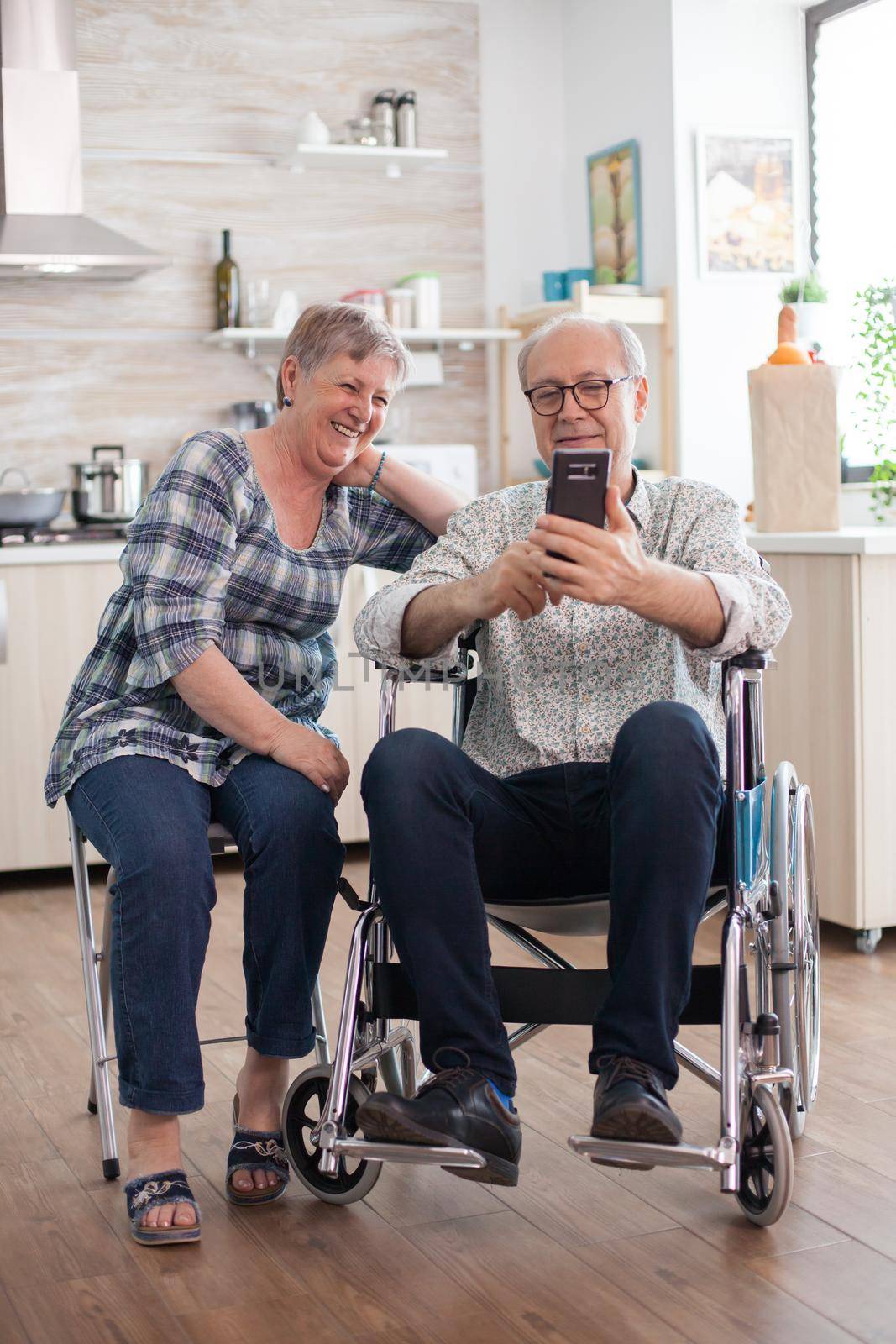 Disabled senior man in wheelchair and his wife Laughing and browsing on modern smartphone in kitchen. Paralyzed old man and his wife having a online conference.