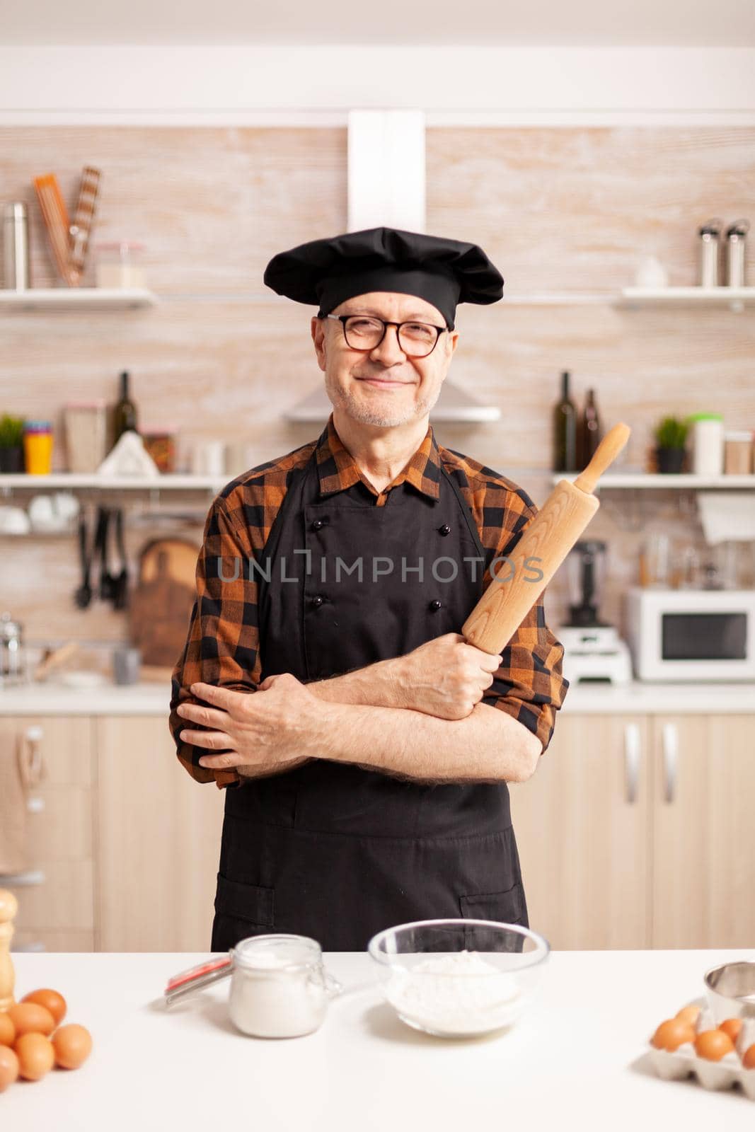 Caucasian old man wearing apron in home kitchen smiling at camera. Retired elderly baker in kitchen uniform preparing pastry ingredients on wooden table ready to cook homemade tasty bread, cakes and pasta