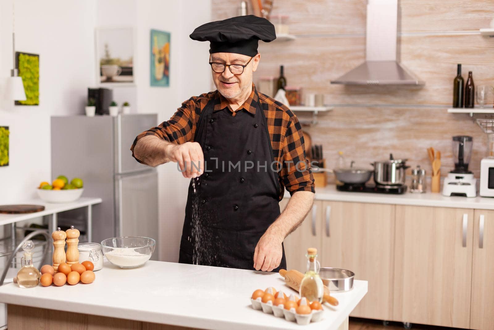 Experienced baker in kitchen preparing delicious pizza using bio wheat flour. Retired senior chef with bonete and apron, in kitchen uniform sprinkling sieving sifting ingredients by hand.