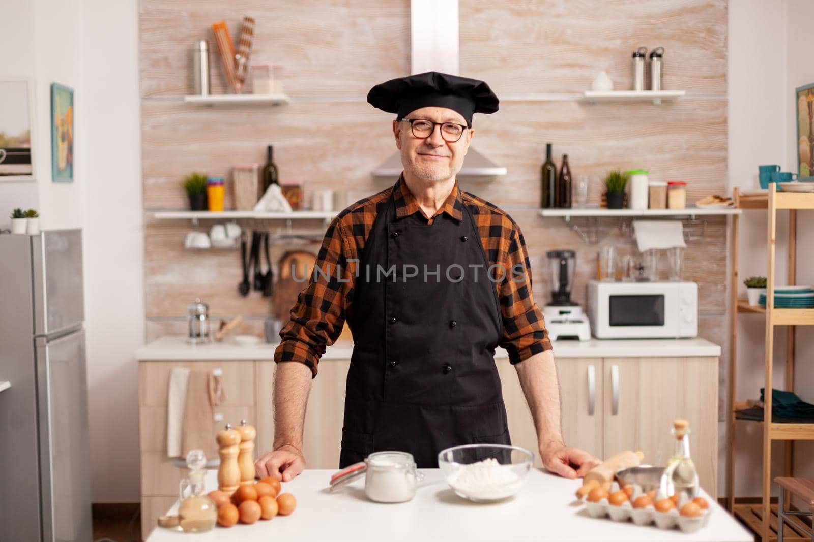 Portrait of chef wearing bonete looking at camera and smiling. Retired elderly baker in kitchen uniform preparing pastry ingredients on wooden table ready to cook homemade tasty bread, cakes and pasta