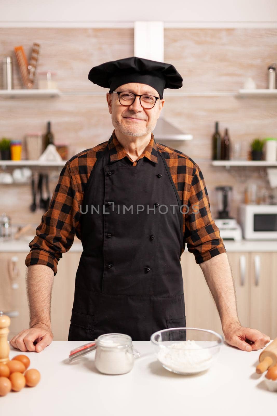 Senior baker smiling looking at camera in kitchen Retired elderly chef in kitchen uniform preparing pastry ingredients on wooden table ready to cook homemade tasty bread, cakes and pasta
