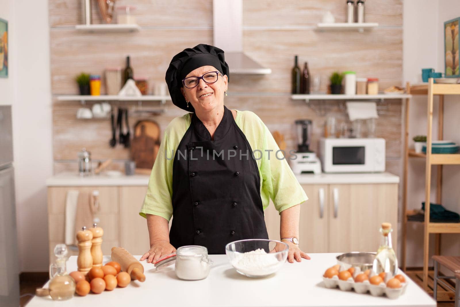 Senior woman wearing baker uniform in home kitchen smiling at camera. Retired elderly baker in kitchen uniform preparing pastry ingredients on wooden table ready to cook homemade tasty bread.