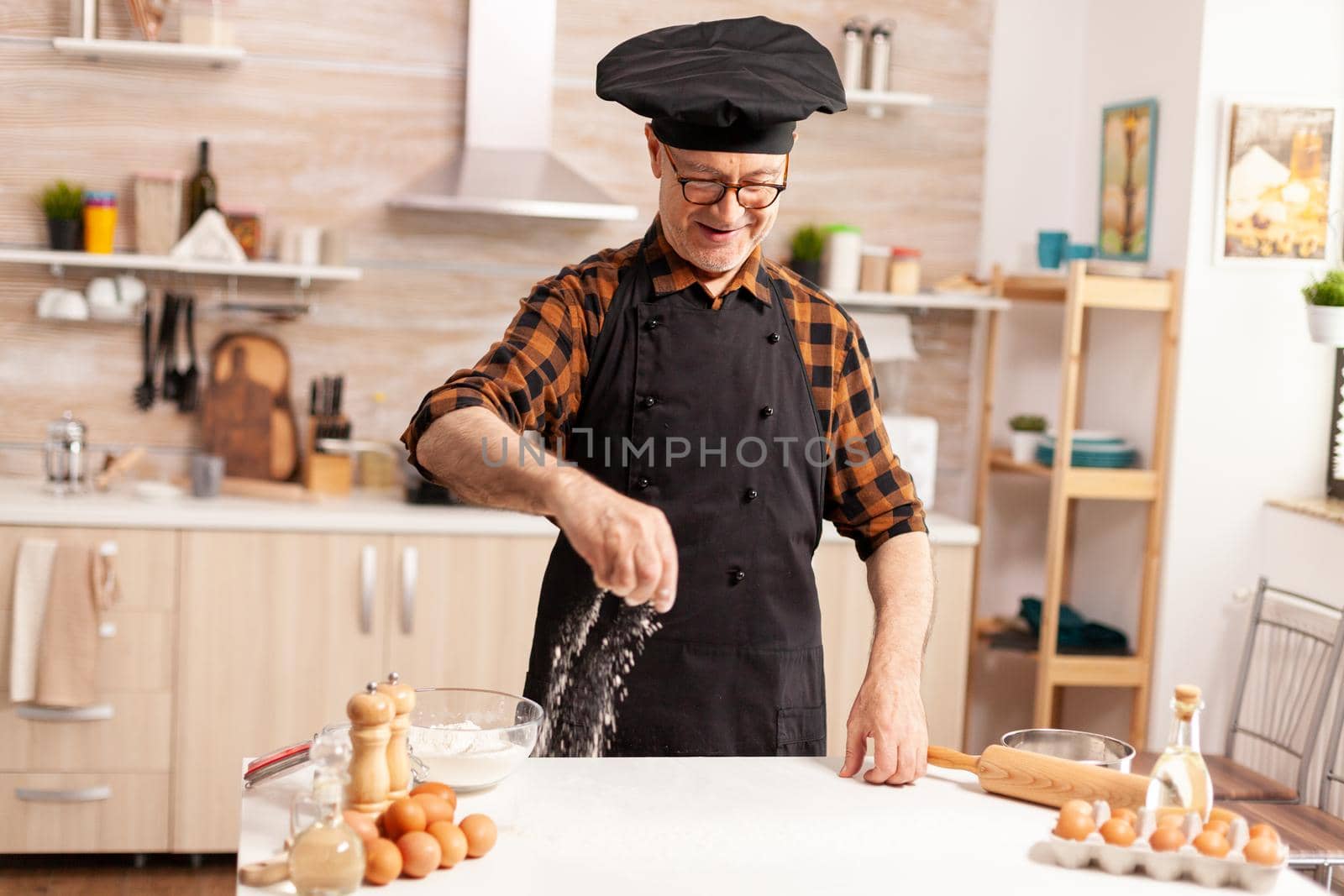 Retired chef in home kitchen wearing apron while sprinkling ingredient on table for tasty. Retired senior chef with bonete and apron, in kitchen uniform sprinkling sieving sifting ingredients by hand.