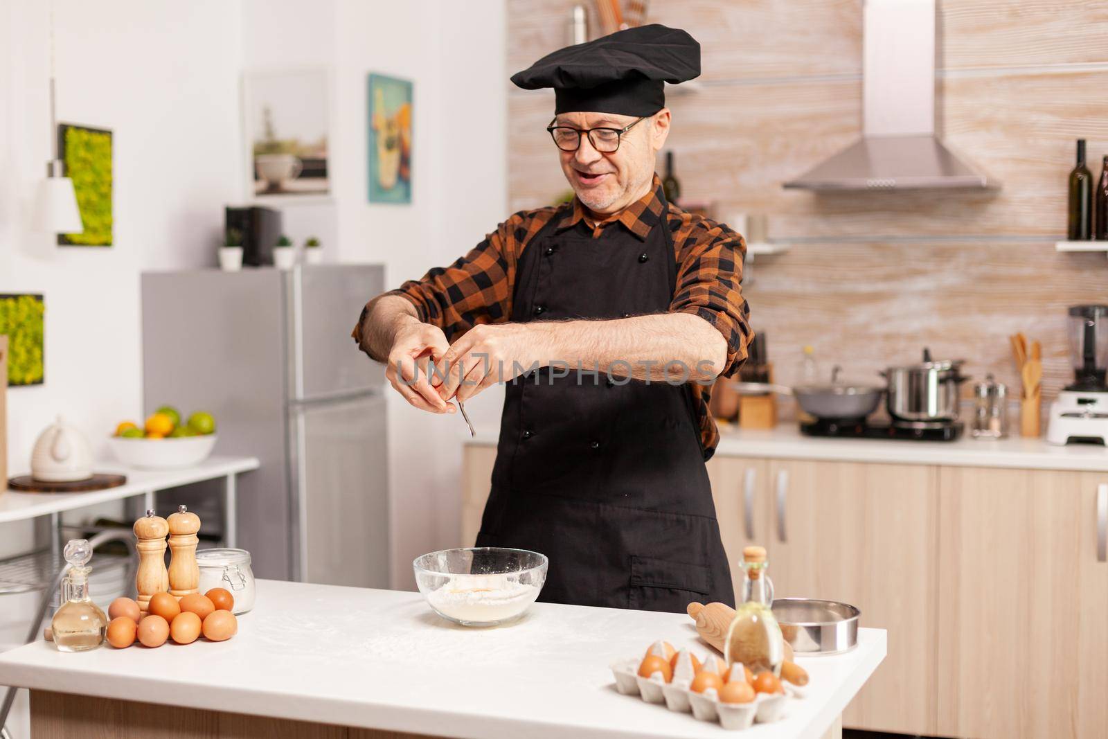 Elderly man wearing apron and bonete preparing tasty pizza cracking eggs on flour. Elderly pastry chef cracking egg on glass bowl for cake recipe in kitchen, mixing by hand, kneading.