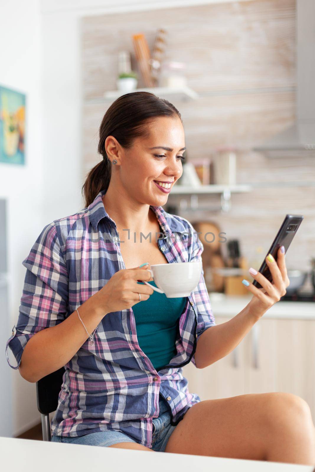 Woman smiling using smartphone and enjoying a cup of hot green tea in kitchen. Holding phone device with touchscreen using internet technology scrolling, searching on intelligent gadget.
