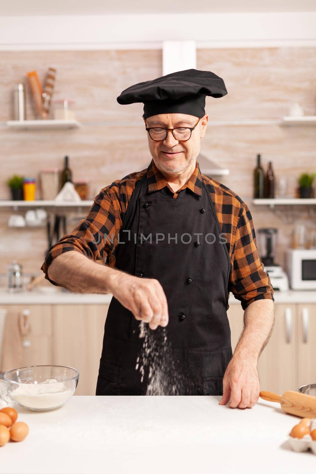 Happy retired chef preparing homemade pizza using bio wheat flour. Retired senior chef with bonete and apron, in kitchen uniform sprinkling sieving sifting ingredients by hand.