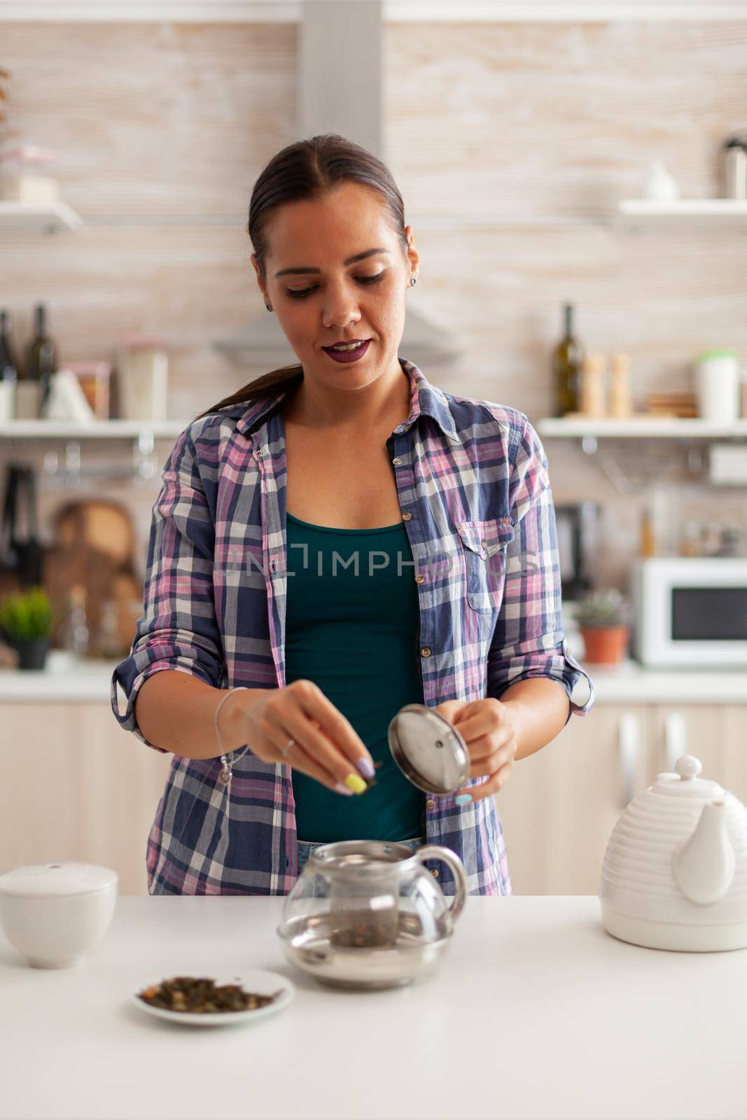 Using natural herbs in kitchen to prepare tea during breakfast. Preparing tea in the morning, in a modern kitchen sitting near the table. Putting with hands, healthy herbal in pot.