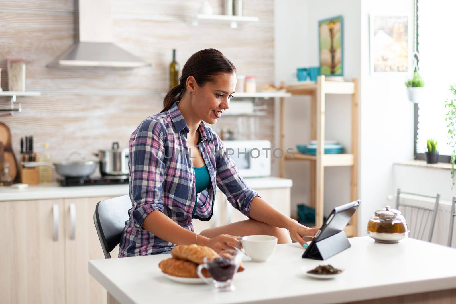 Woman browsing on tablet pc during breakfast in kitchen and holding cup of green tea. Working from home using device with internet technology, typing, on gadget during breakfast.