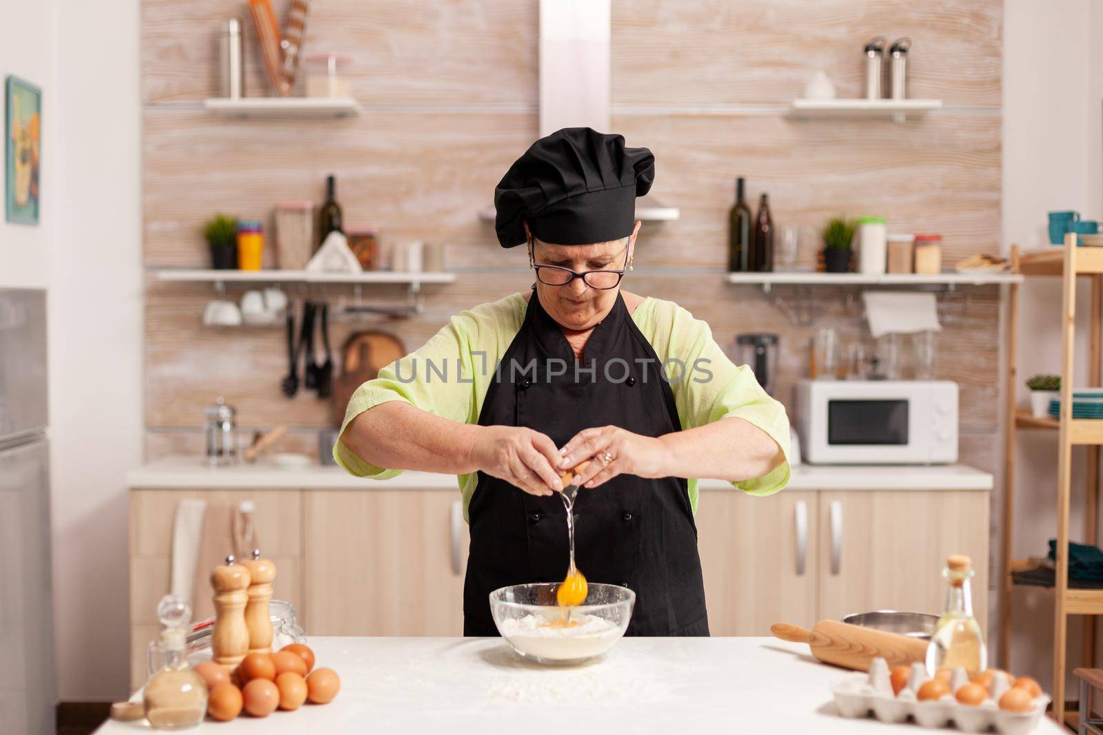Elderly woman cracking eggs over wheat flour in home kitchen. Elderly pastry chef cracking egg on glass bowl for cake recipe in kitchen, mixing by hand, kneading