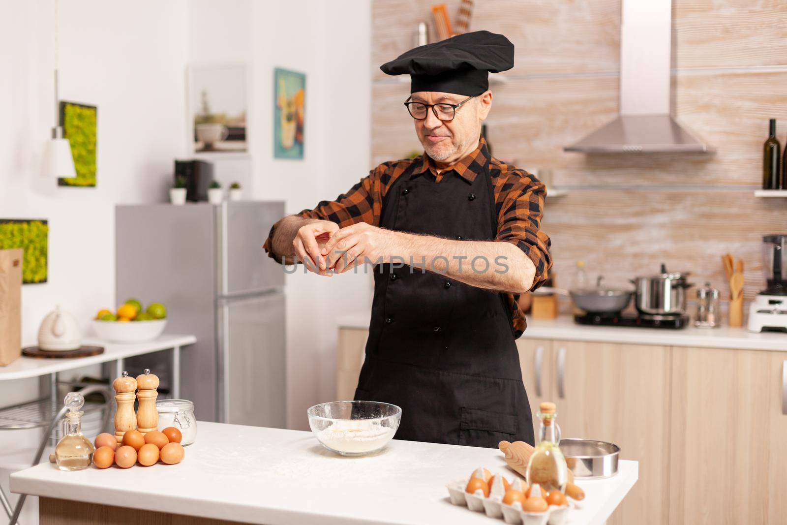 Elderly man cracking eggs over wheat flour while preparing delicious food. Elderly pastry chef cracking egg on glass bowl for cake recipe in kitchen, mixing by hand, kneading ingredients prreparing homemade cake