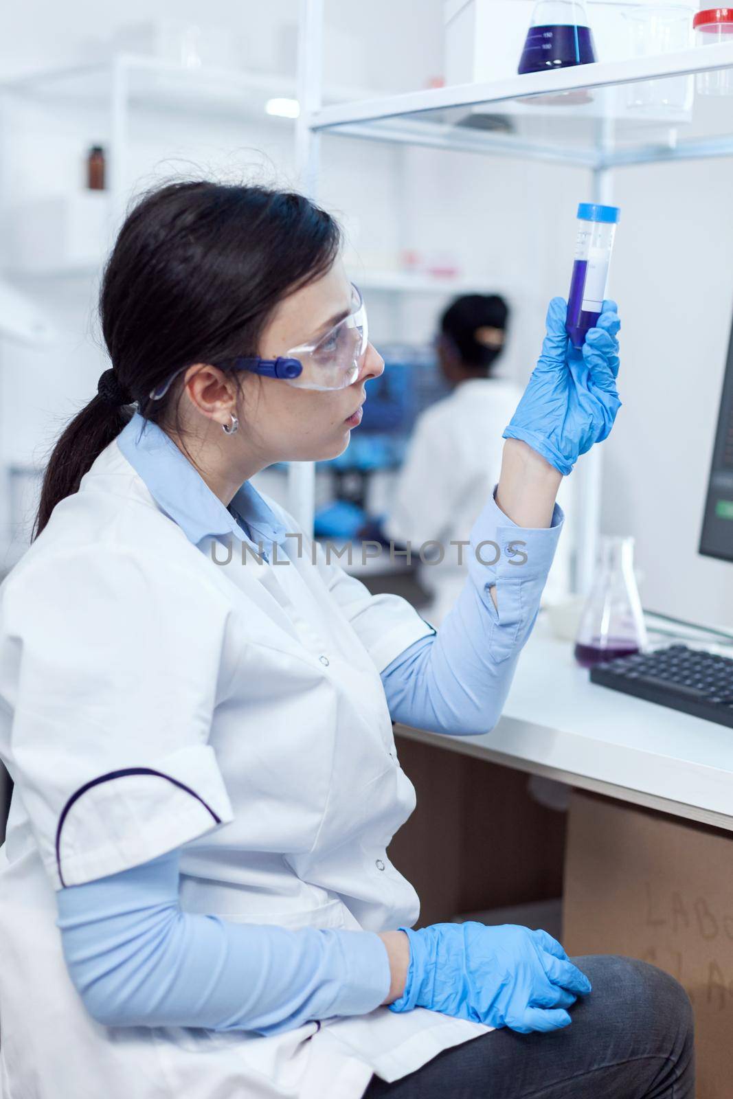 Young scientist analysing scientific sample on plastic test tube with african coworker sitting at her workplace. Chemistry researcher in sterile laboratory using modern technology to test microbiology liquid.