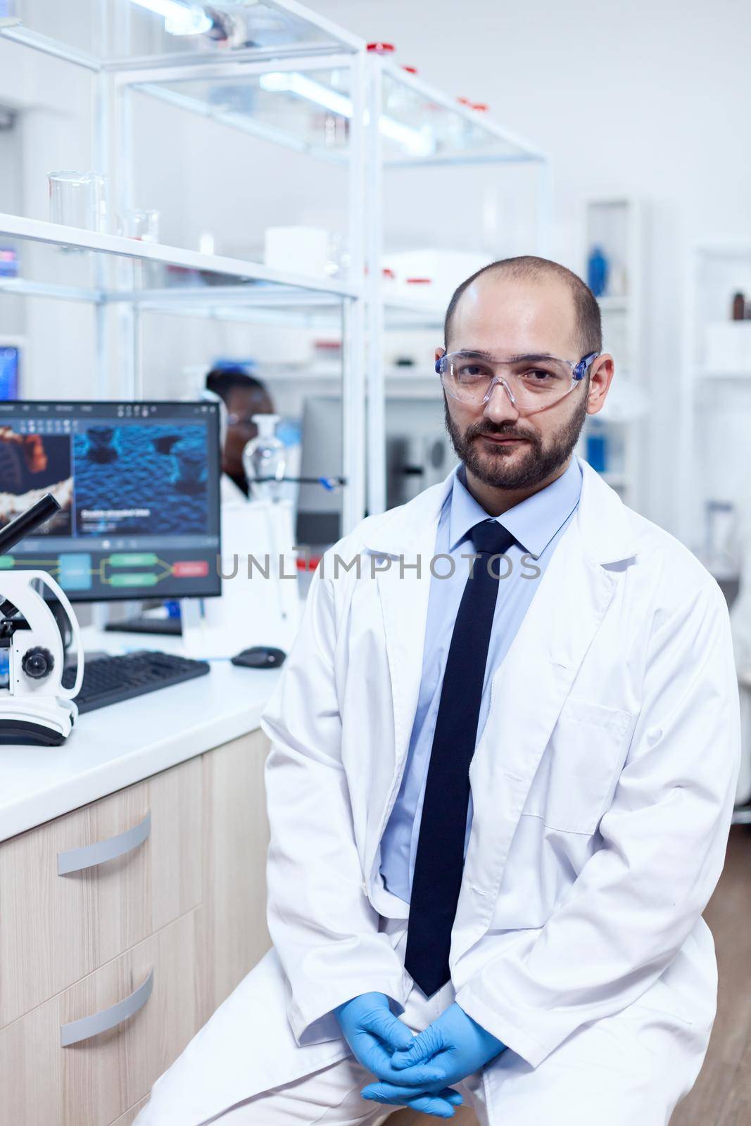 Healthcare researcher wearing lab coat and protective glasses looking at camera. Serious expert in genetics in lab with modern technology for medical investigations with african assistant in the background.