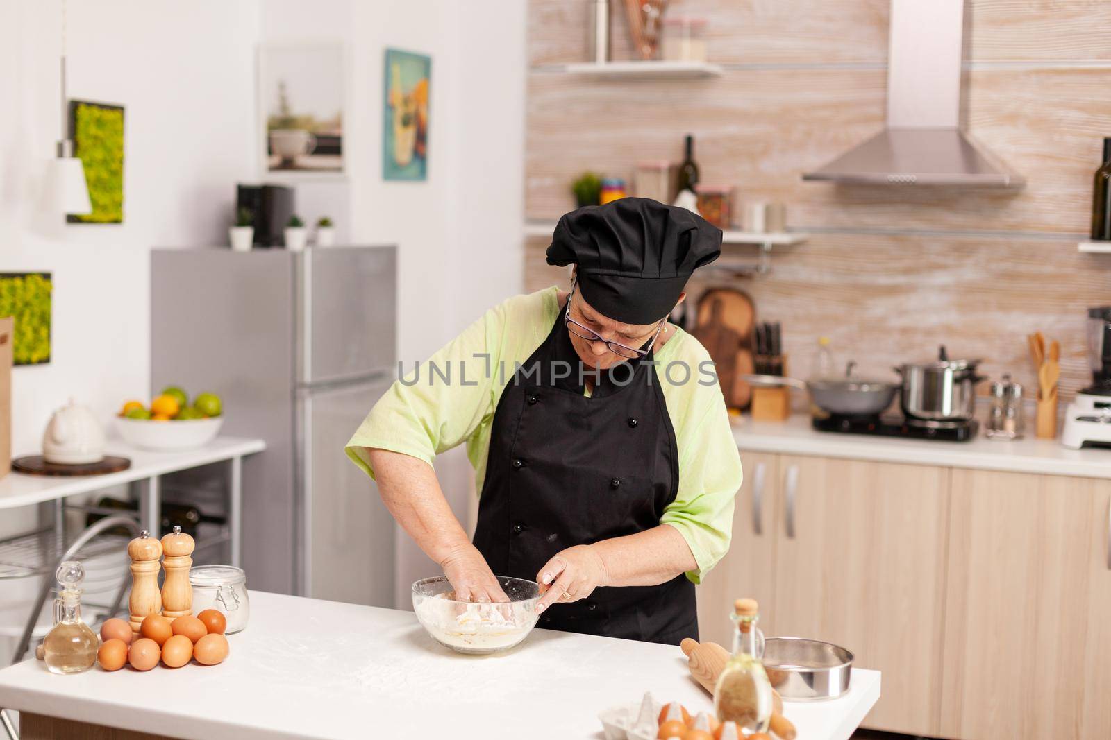 Elderly woman wearing bonete while preparing dough for paste in home kitchen following traditional recipe. Retired elderly chef with uniform sprinkling, sieving sifting raw ingredients and mixing.