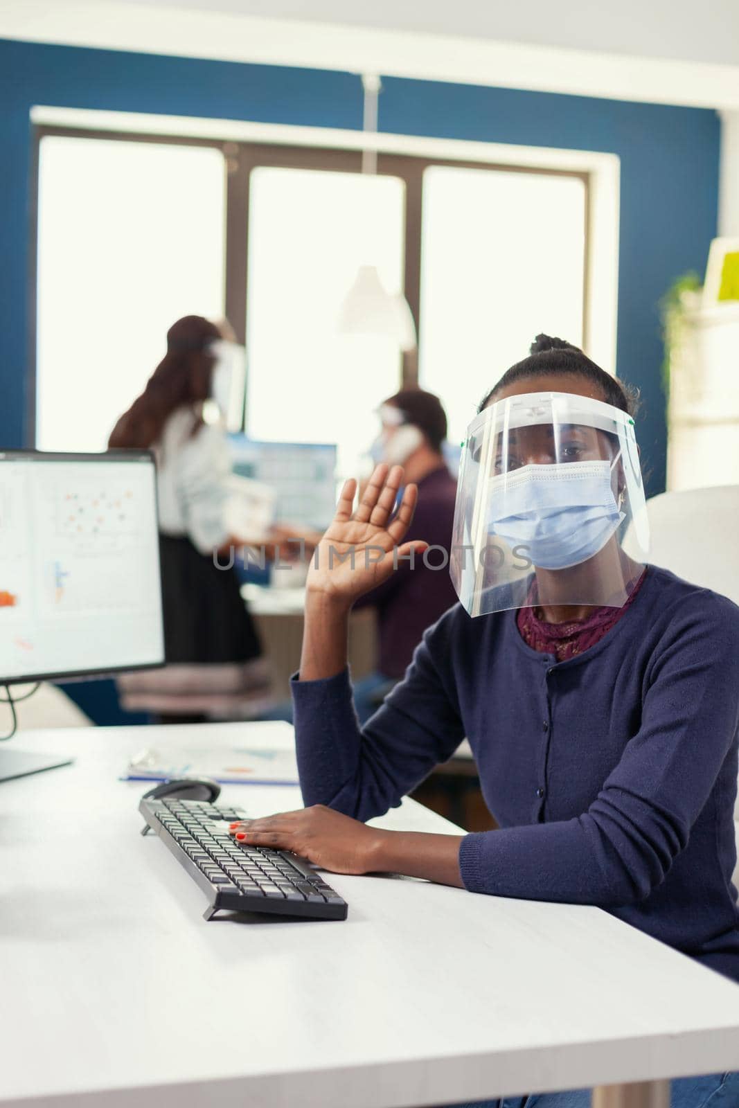 Pov of african worker in remotely call in workplace sitting at desk wearing face mask against covid19. Woman speaking with team during online conference while colleagues working in background.