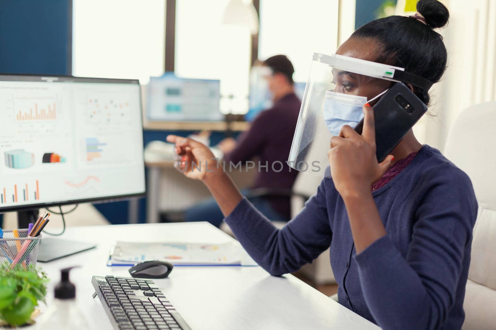 African freelancer discussing on smartphone about financial graphs wearing face mask during coronavirus. Multiethnic coworkers working respecting social distance in financial company.