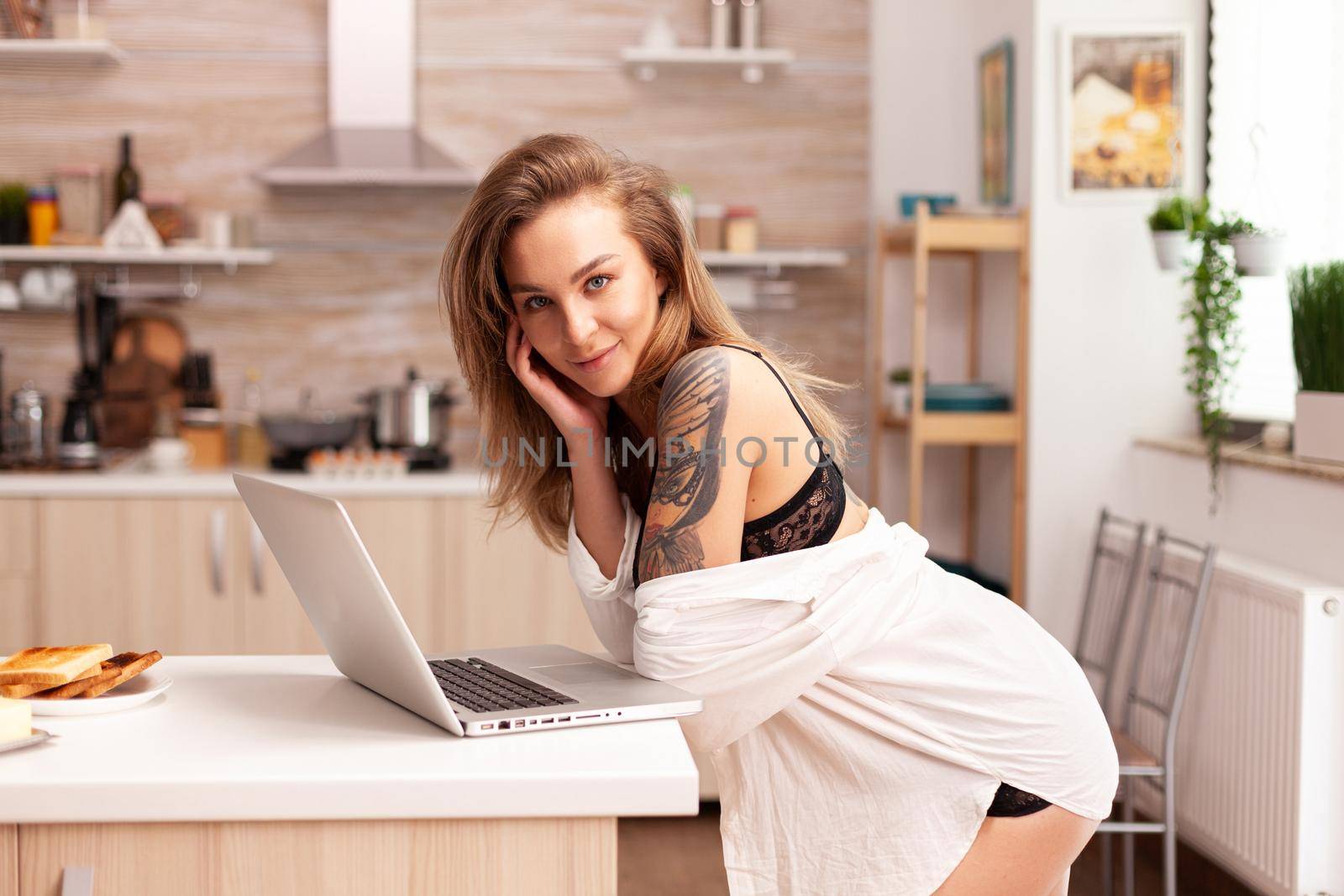 Care free woman during breakfast wearing sexy underwear using laptop. Attractive blonde lady with tattoos typing on pc sitting in the kitchen dressed in seductive underwear smiling.