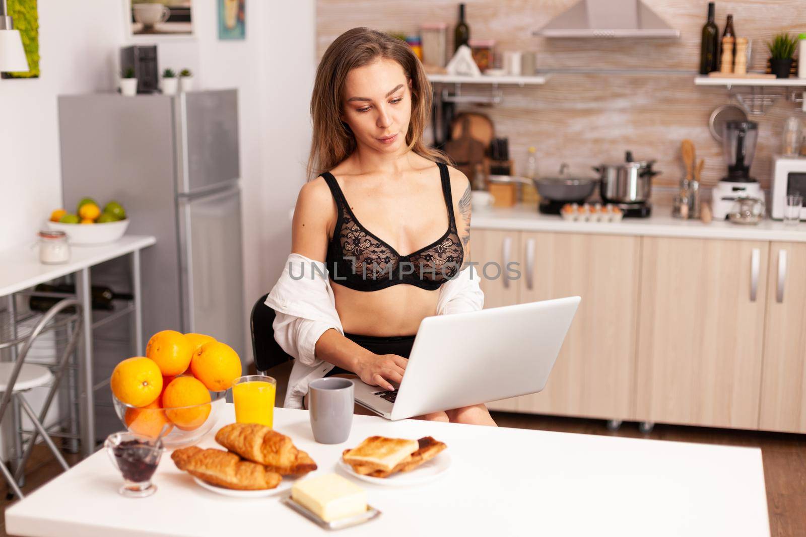 Attractive lady with tattoos in sexy lingerie searching on laptop in home kitchen. Attractive blonde lady with tattoos typing on pc sitting in the kitchen dressed in seductive underwear smiling