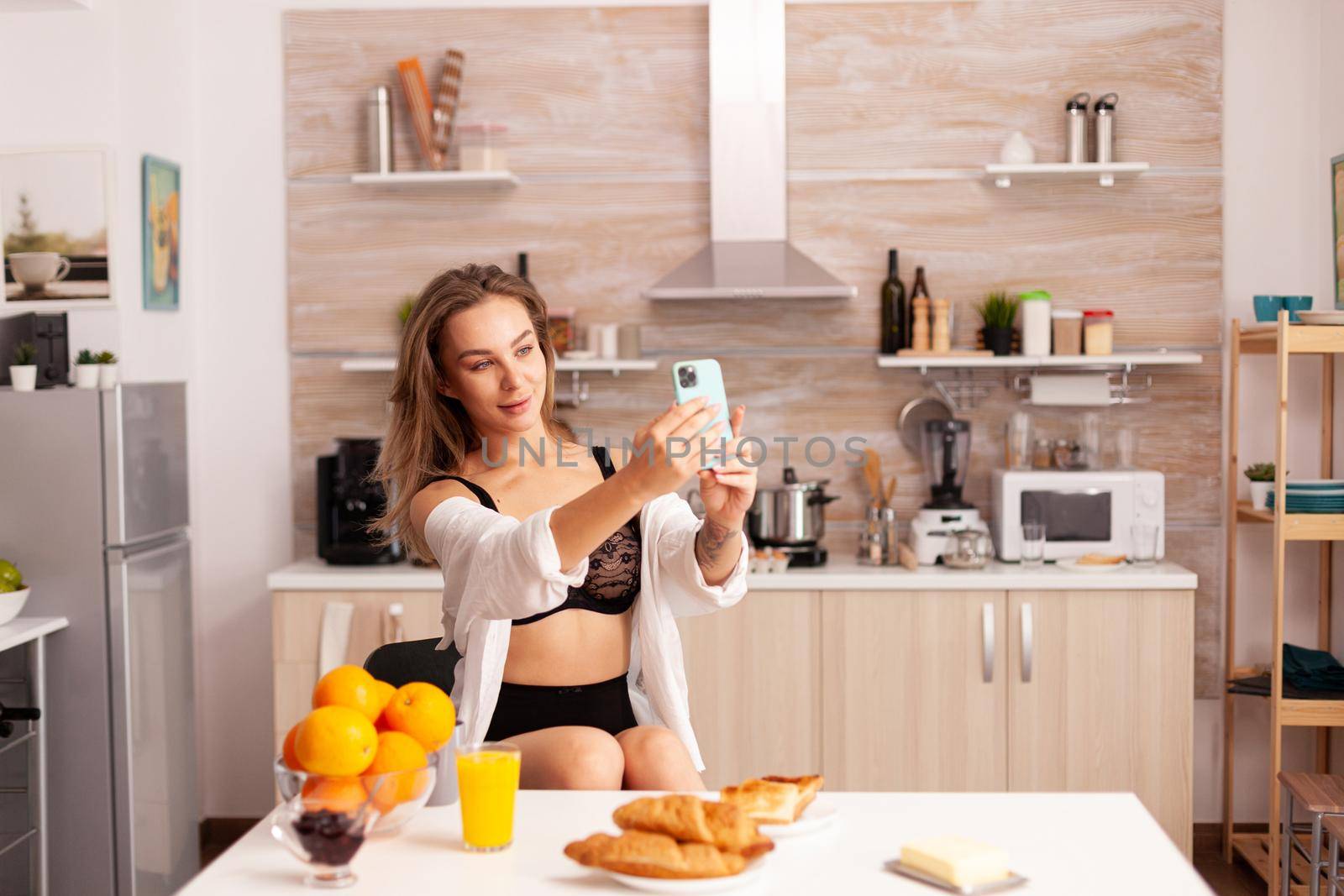 Seductive woman in sexy lingerie taking selfie using smartphone in home kitchen. Attractive lady with tattoos using smartphone wearing temping underwear in the morning.