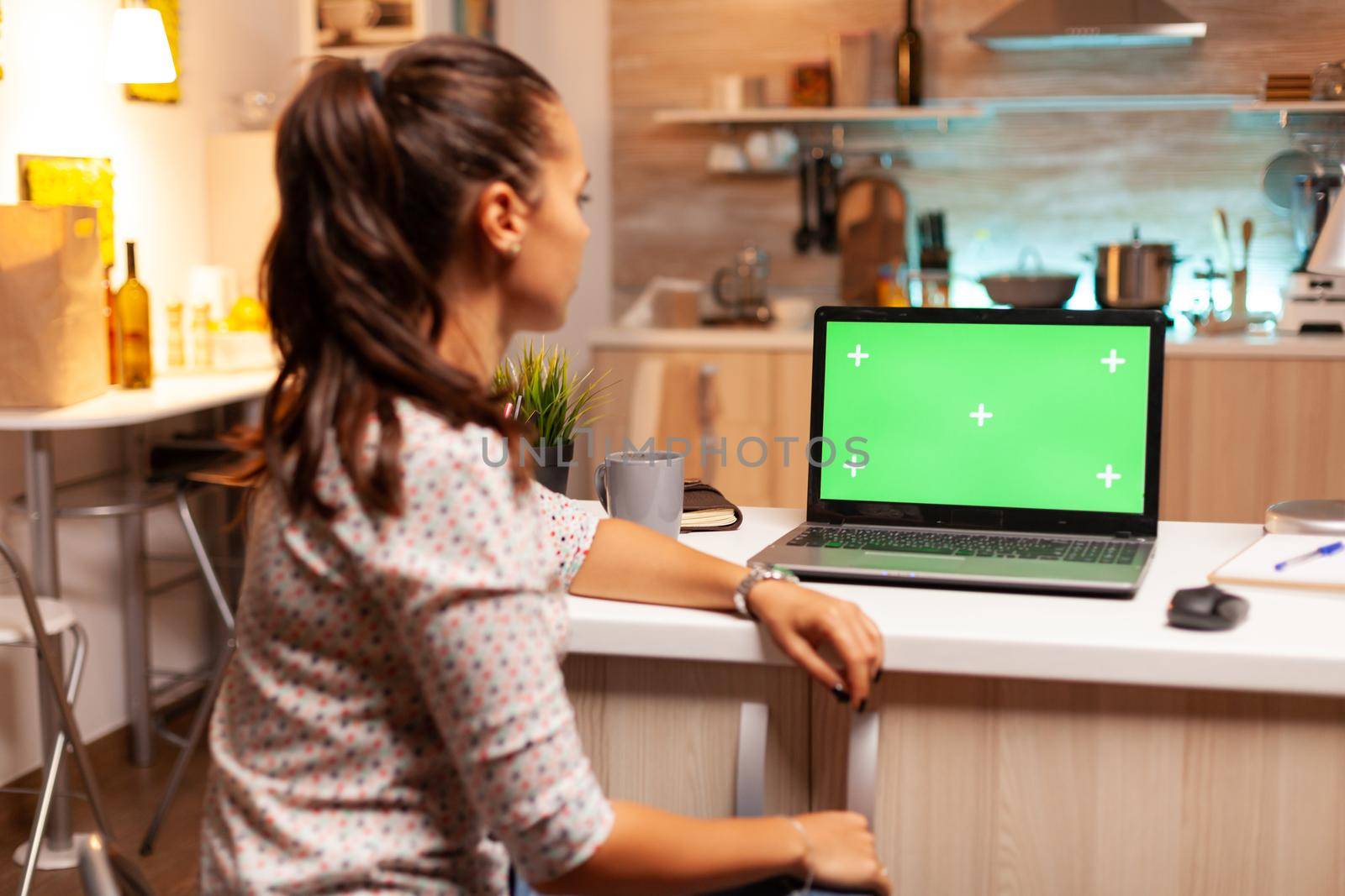 Woman looking at laptop with green mockup during night time in home kitchen. Sitting at desk works on computer late at night, business, online, smart, blank, copyspace.