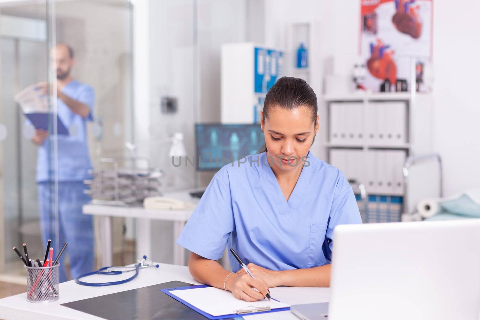 Healhcare practitioner checking patient insurance on laptop and taking notes. Health care physician using computer in modern clinic looking at monitor, medicine, profession, scrubs.