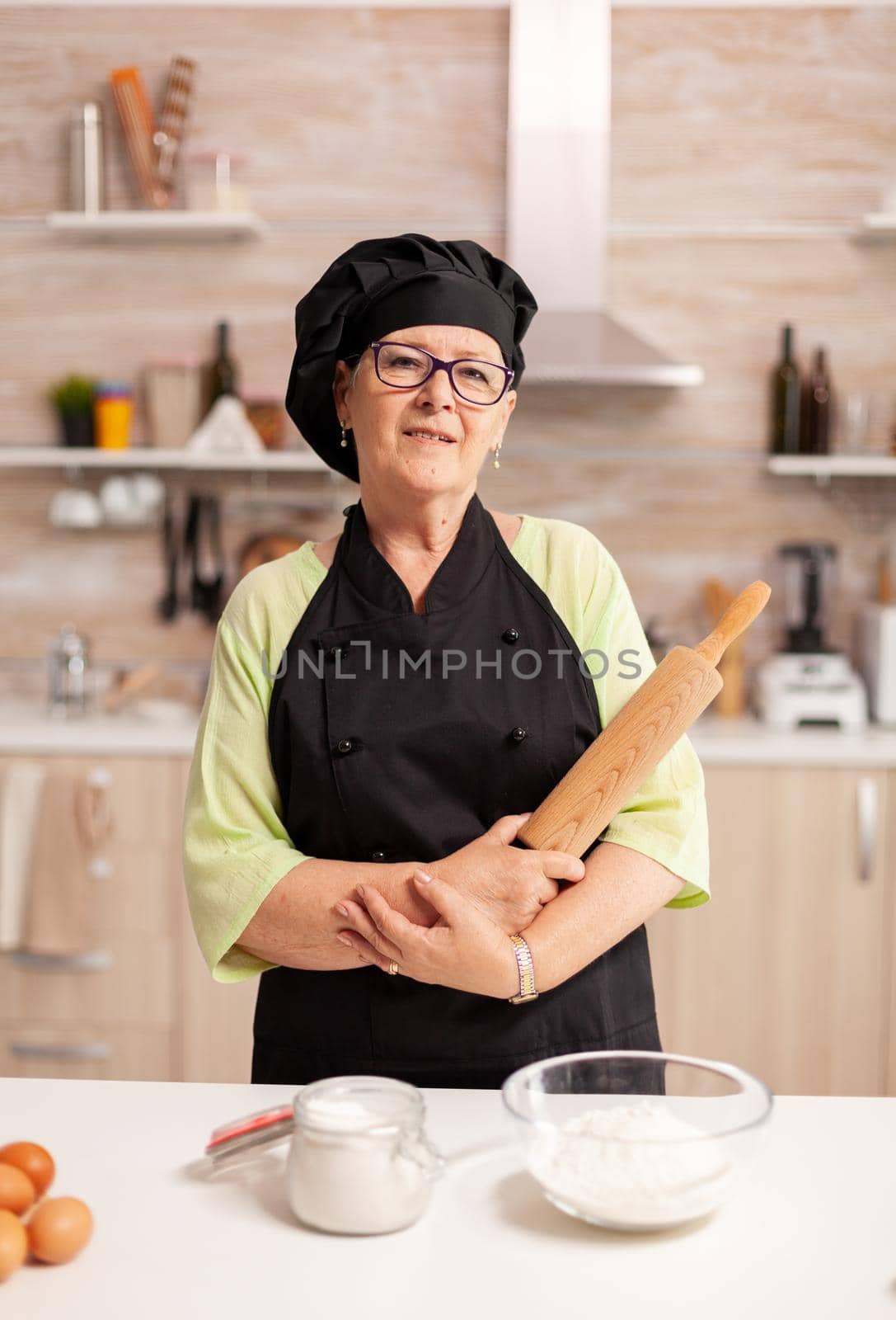 Professional baker smiling at camera in home kitchen wearing apron and bonete. Retired elderly baker in kitchen uniform preparing pastry ingredients on wooden table ready to cook homemade tasty bread,