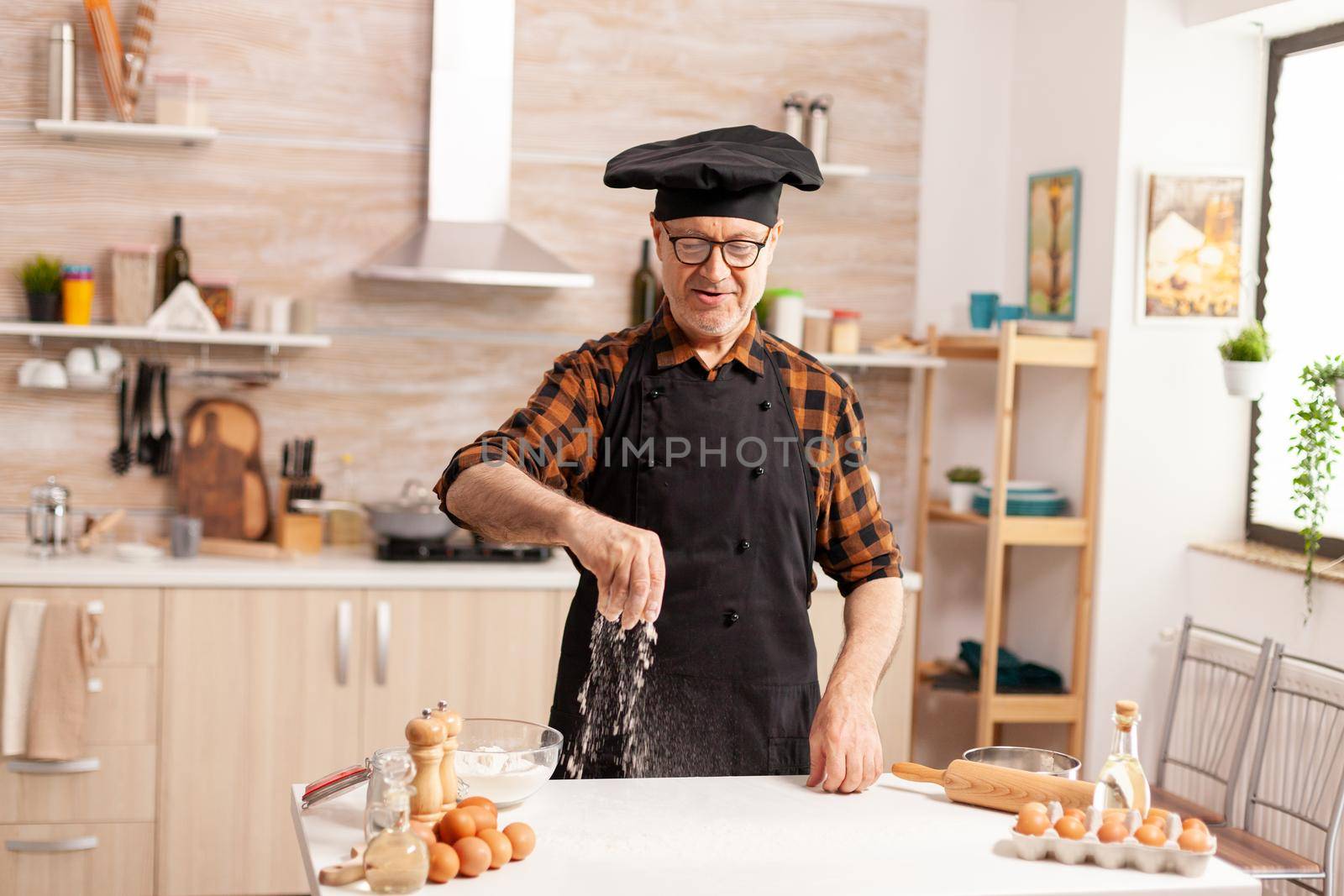 Elderly baker preparing tasty food using bio wheat flour wearing apron and bonete. Retired senior chef with bonete and apron, in kitchen uniform sprinkling sieving sifting ingredients by hand.