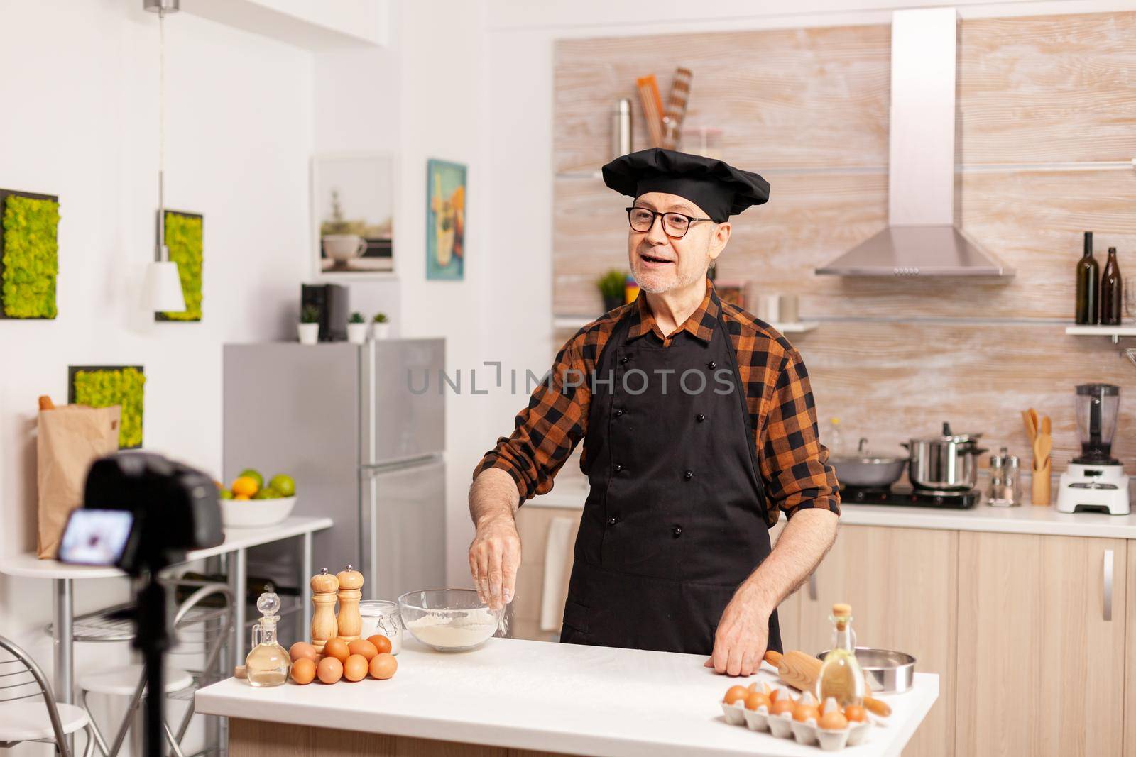 Baker recording for podcast in kitchen while sifting flour on table. Retired blogger baker influencer using internet technology communicating, shooting, blogging on social media with digital equipment.