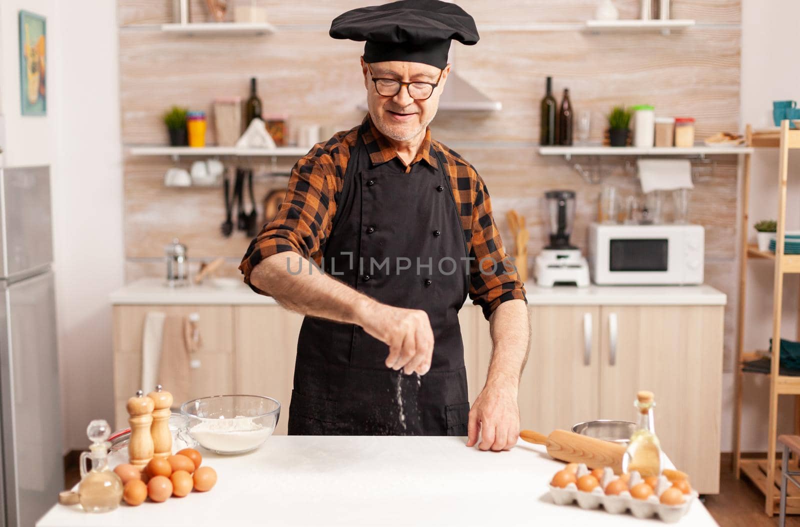 Retired cook preparing tasty food spreading wheat flour on kitchen table top. Retired senior chef with bonete and apron, in kitchen uniform sprinkling sieving sifting ingredients by hand.