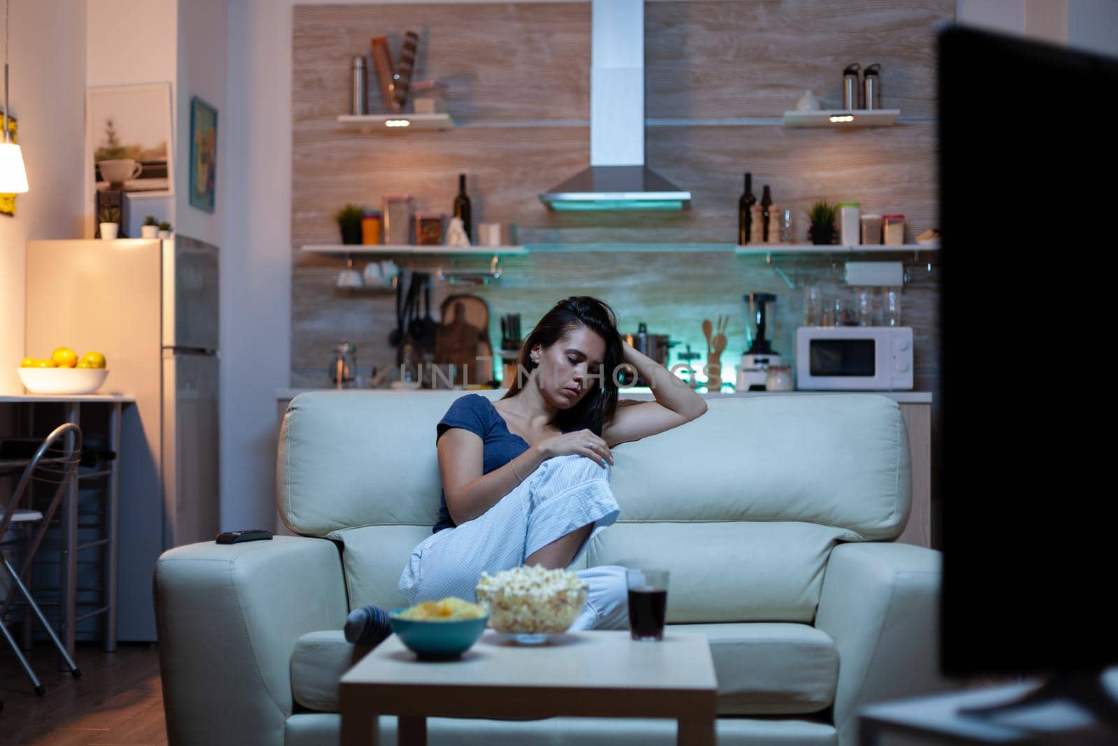 Caucasian lady asleep on sofa alone in living room while watching movie on tv late at home at night. Tired lonely housewife in pijamas falling asleep in front of TV sitting on comfortable couch.