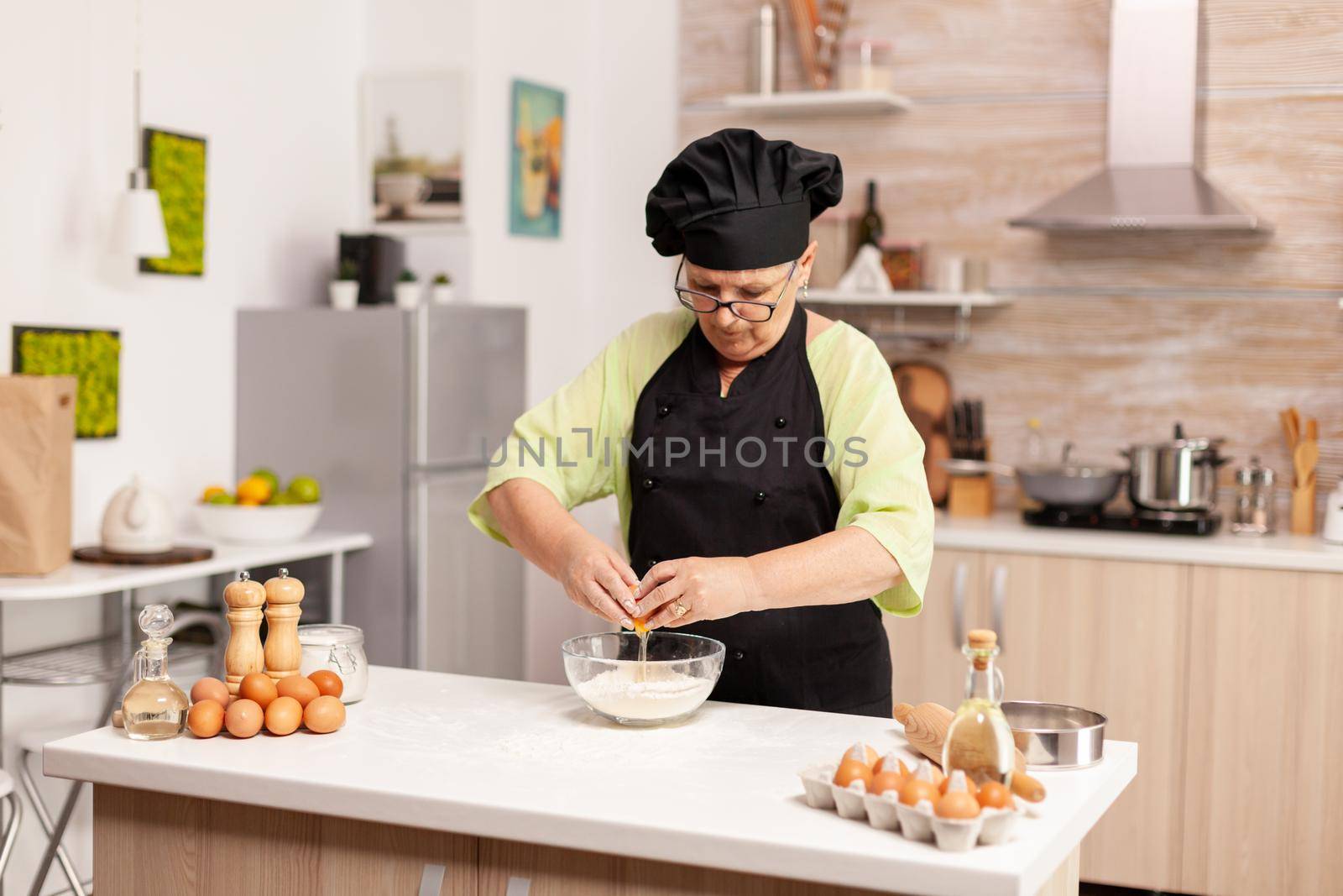 Elderly pastry chef cracking egg on glass bowl for cake recipe in kitchen., Mixing by hand, kneading ingredients prreparing homemade cake