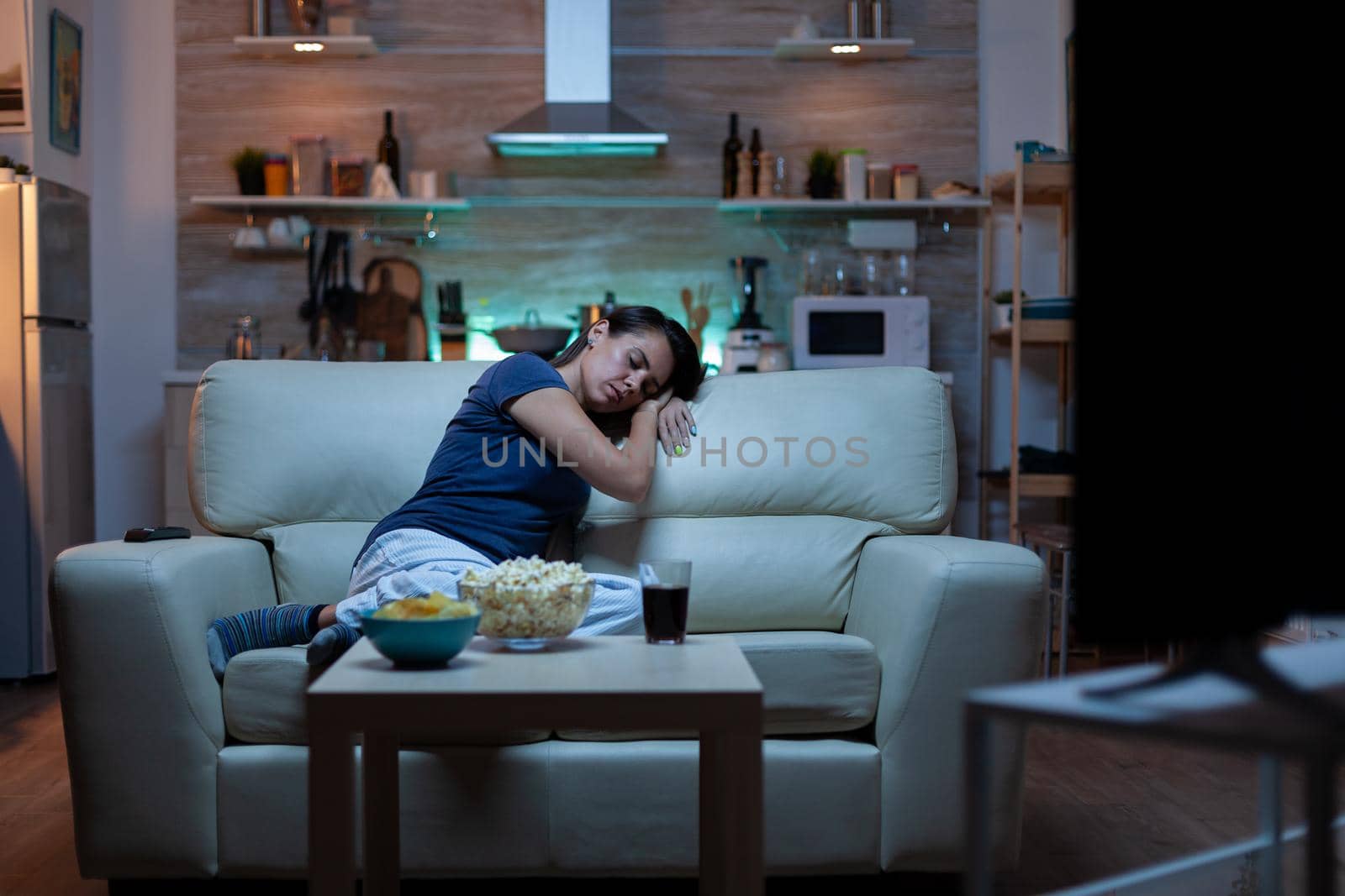 Young tired after work woman looking asleep in the evening in front of TV. Exhausted lonely sleepy lady in pajamas sleeping on sofa while watching a bored movie in living room, closing eyes at night