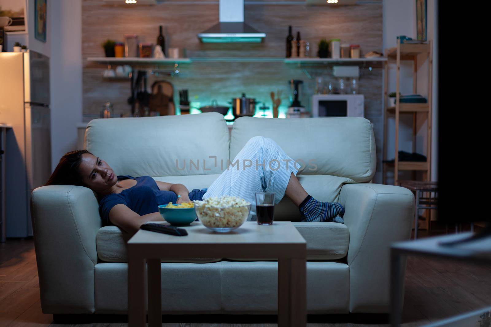 Young woman watching TV and feeling bored sitting on the couch in the living room at home. Tired, exhausted, lonely lady relaxing watching tv lying on comfortable sofa eating snacks late at night