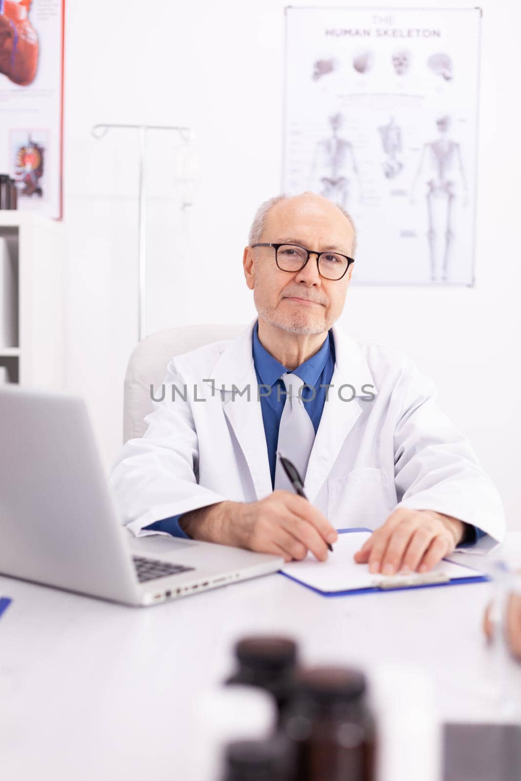 Mature doctor in hospital office wearing lab coat while looking at camera. Medical practitioner using notebook in clinic workplace , confident, expertise, medicine.