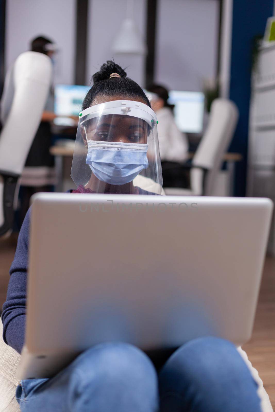 African businesswoman working on laptop wearing face mask against covid19. Multiethnic business team working respecting social distance during global pandemic with coronavirus.