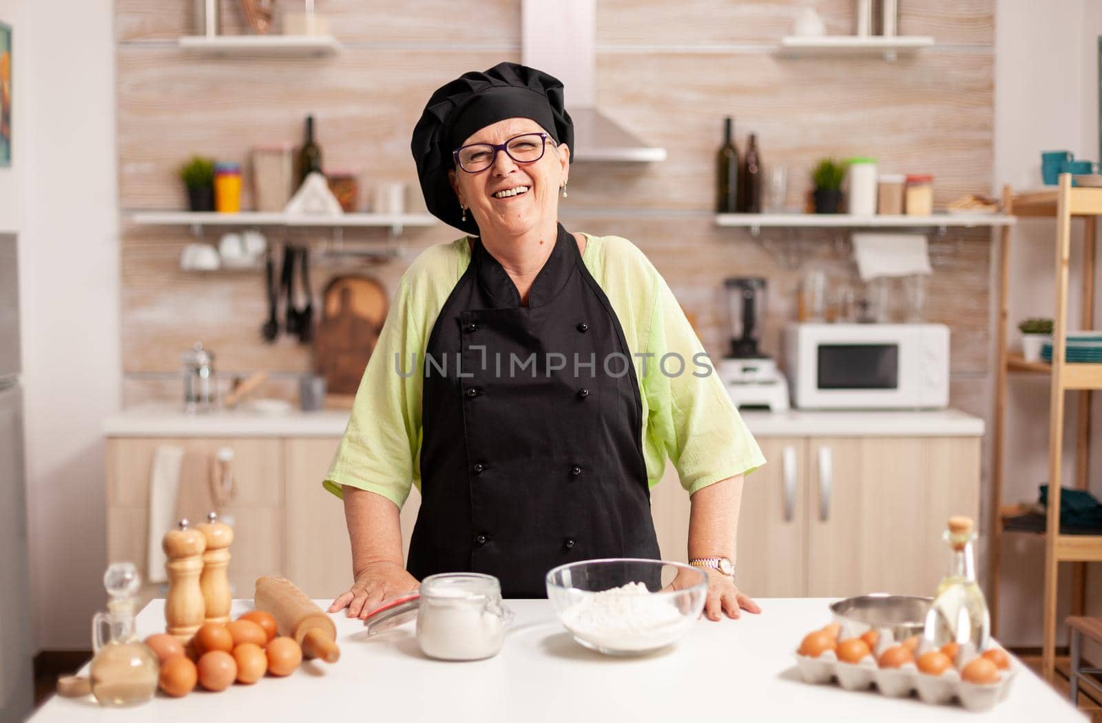 Happy elderly woman wearing bonete looking at camera in dining room at home. Retired elderly baker in kitchen uniform preparing pastry ingredients on wooden table ready to cook homemade tasty bread.