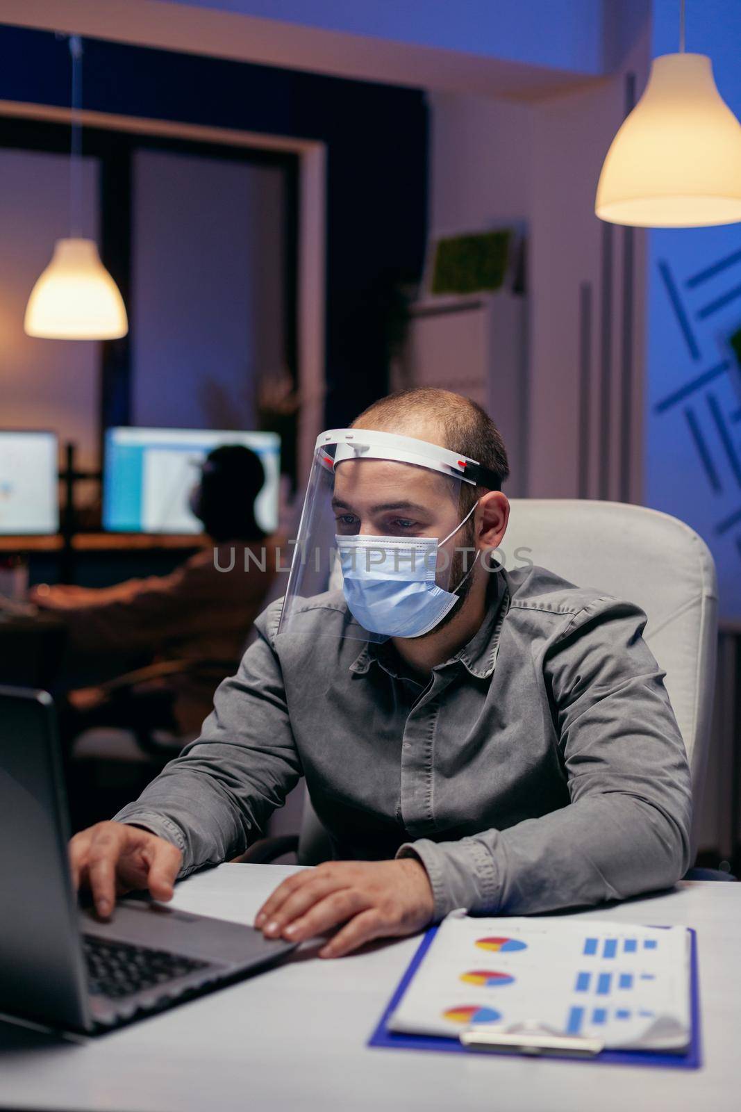 Manager and colleagues doing overtime with visor against covid-19. Stressed man in corporation working hard to finish a project wearing face mask as safety precaution due to coronavirus pandemic.
