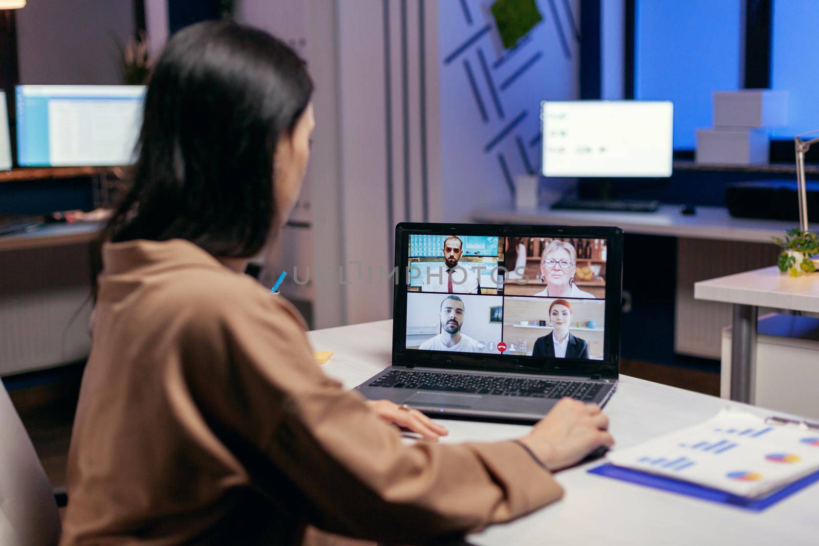 Entrepreneur talking in the course of online meeting with colleagues doing overtime. Woman working on finance during a video conference with coworkers at night hours in the office.