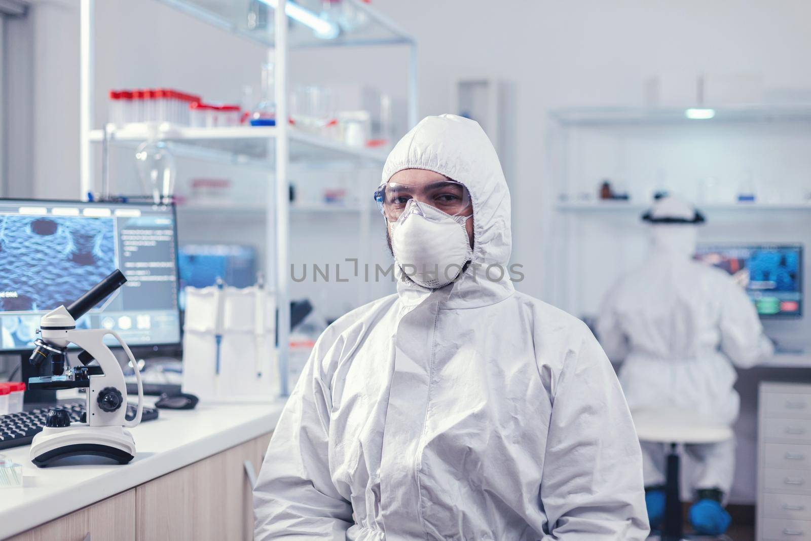 Chemist feeling tired from work for a long time in laboratory sitting at workplace. Overworked researcher dressed in protective suit against invection with coronavirus during global epidemic.