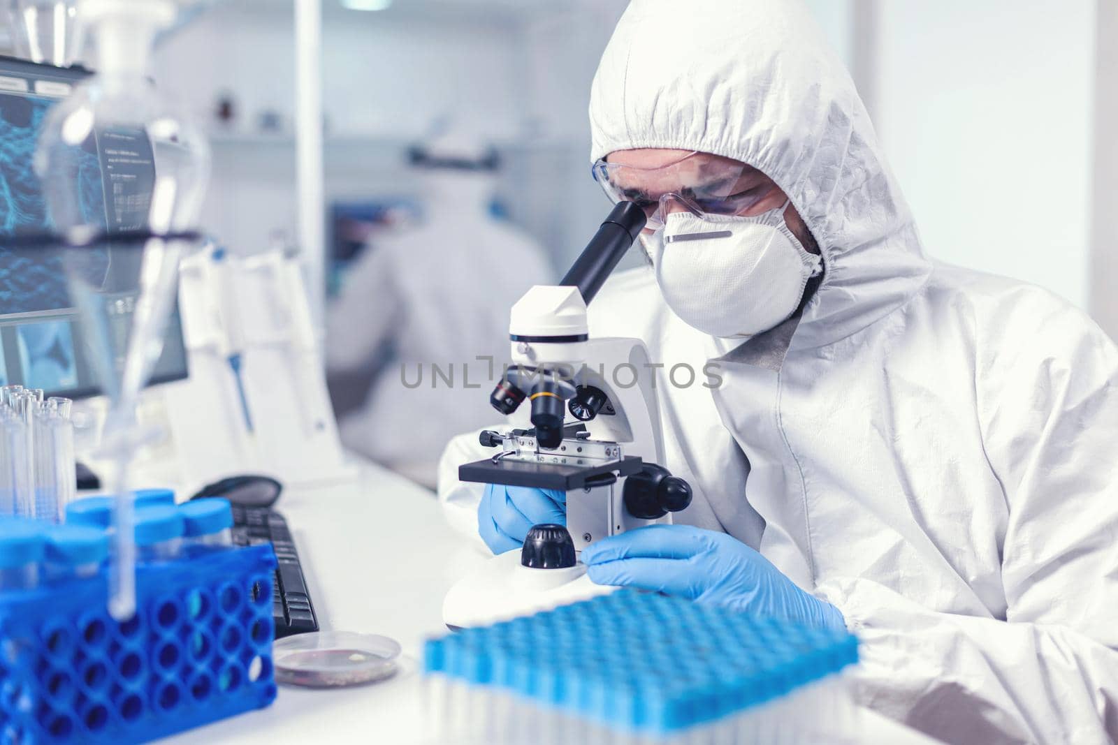 Focused healthcare researcher with face mask and ppe analysing covid using microscope. Scientist in protective suit sitting at workplace using modern medical technology during global epidemic.