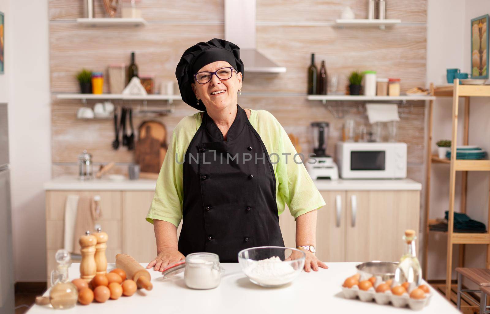 Old lady wearing chef apron and bonete in home kitchen with flour on table. Retired elderly baker in kitchen uniform preparing pastry ingredients on wooden table ready to cook homemade tasty bread.
