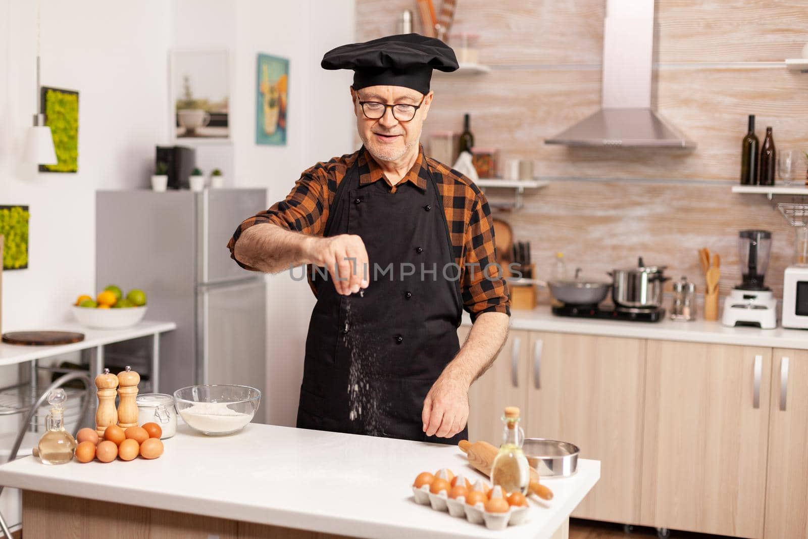 Senior baker sprinkling whear flour on home kitche table for tasty recipe. Retired chef with bonete and apron, in kitchen uniform sprinkling sieving sifting ingredients by hand.